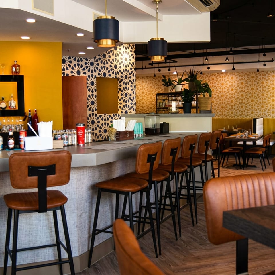 Bright modern restaurant interior with a stocked bar, leather bar stools, patterned walls, pendant lights, and dining tables.