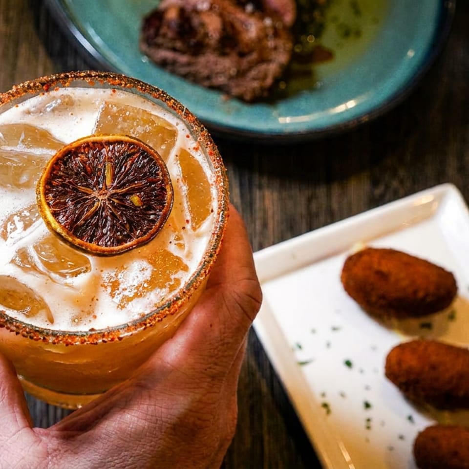Hand holding a cocktail with a dried orange slice, next to croquettes and steak.