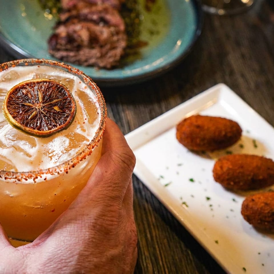 A hand holds a cocktail with a dried orange slice, next to a plate of croquettes.