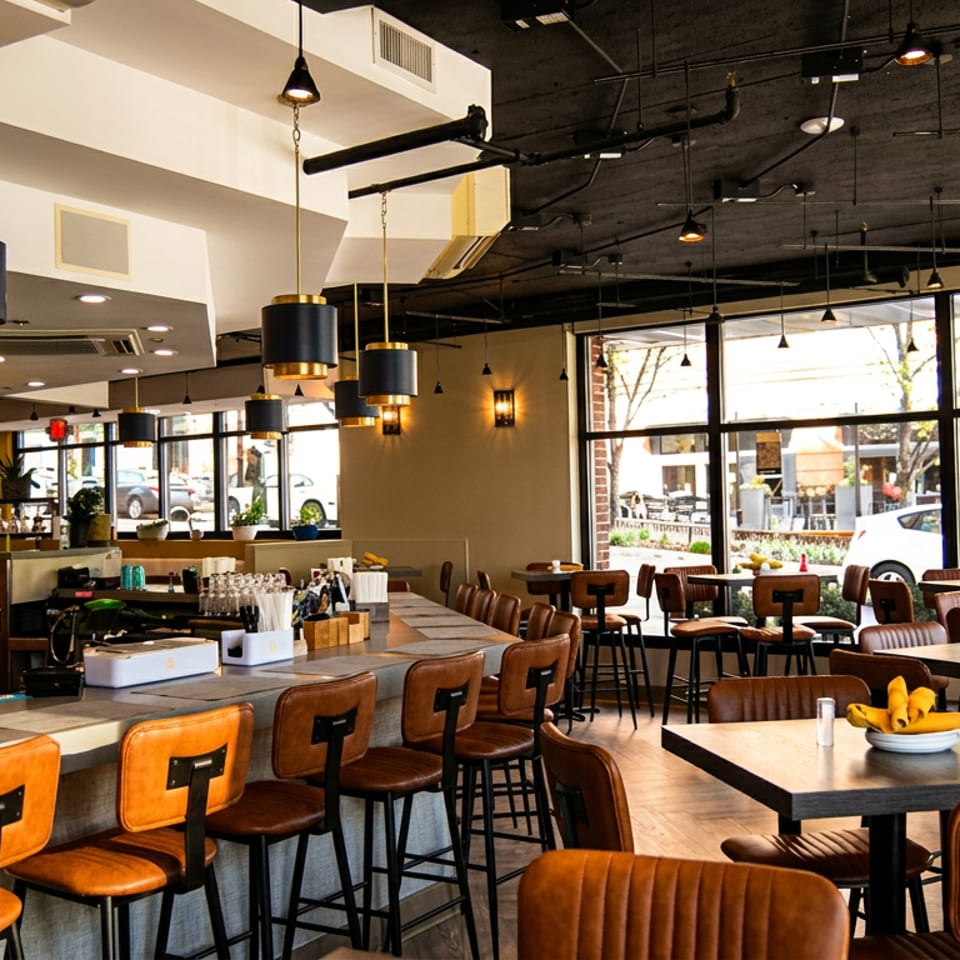 Modern restaurant interior with a long bar, high-top tables, and large windows overlooking a street.