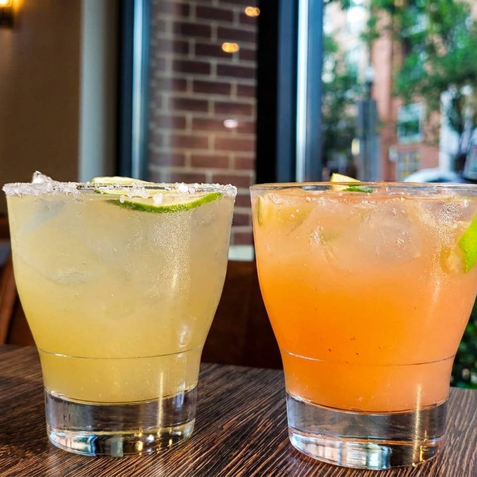 Two cocktails, one yellow and one orange, on a wooden table with a city street in the background.