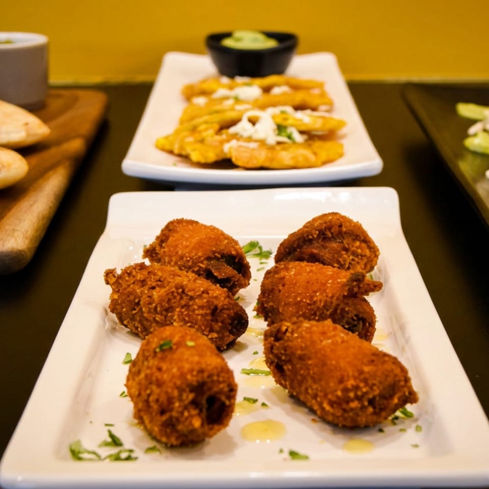Assortment of Latin American appetizers: fried plantains, arepas, stuffed pastries, and croquettes on plates.